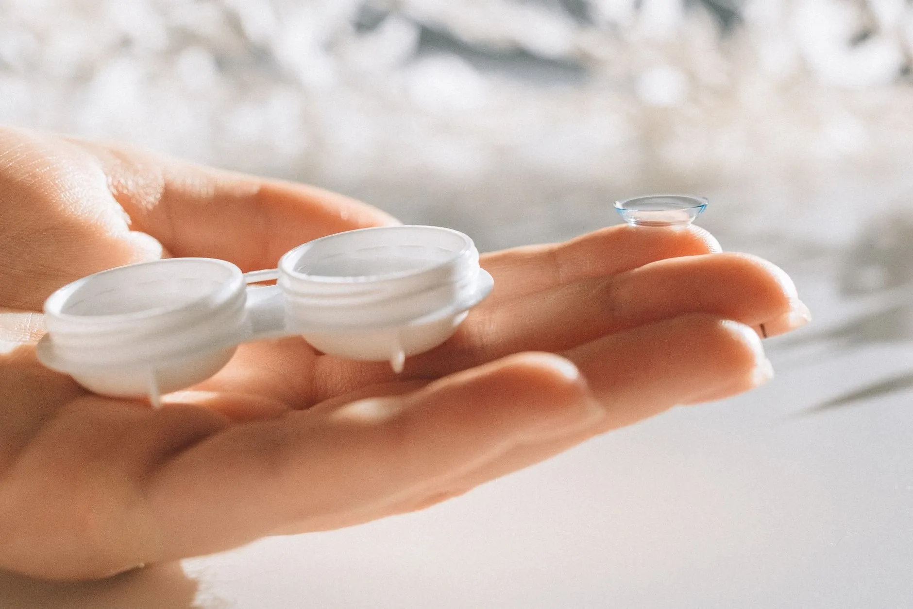 An open contact lens case held in the palm, with a soft lens balanced on a fingertip